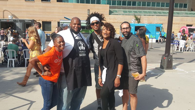Sammy McDowell wearing a white T-shirt and a black apron standing with four friends outside on a sidewalk with food trucks and people walking in the background. 