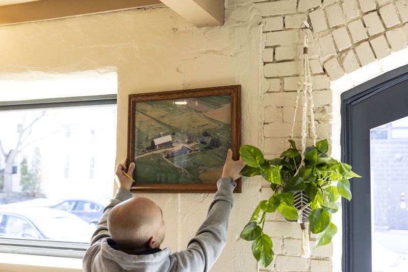 A person in a grey sweatshirt hanging a large framed photo of a farm on a white-washed wall. 