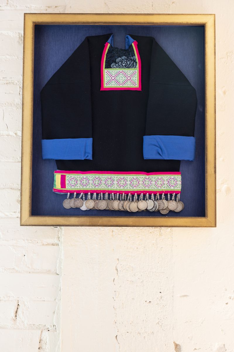 Framed traditional Hmong baby clothing, a black tunic with blue sleeve trimming and an embroidered band at the bottom with rows of attached silver discs hanging from it.