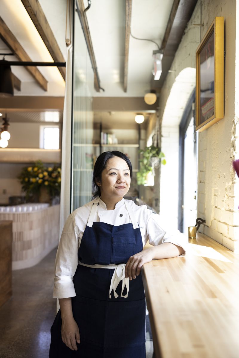 Diane Moua wearing a white shirt and a dark blue apron, standing at the front counter of her restaurant with her elbow up, looking out the window. 