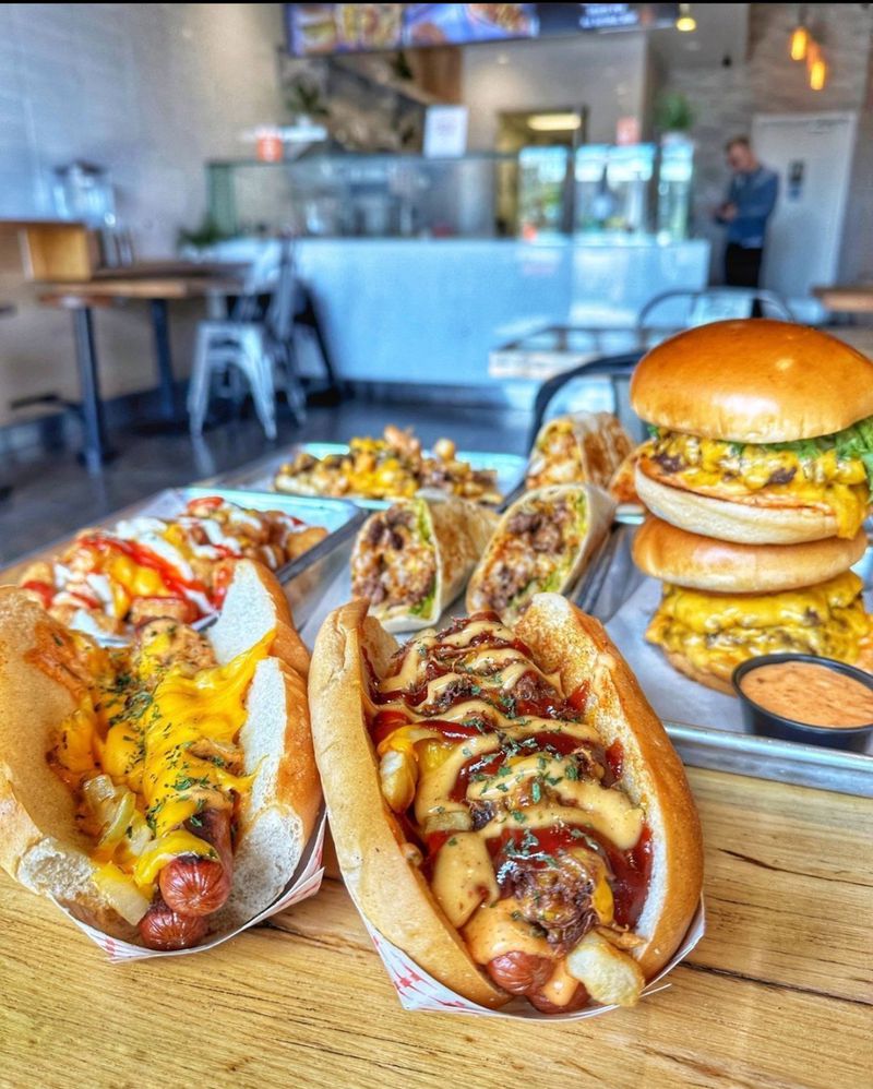 A wooden table set with hot dogs, burgers, wraps, and loaded fries, with a restaurant counter visible in the background. 