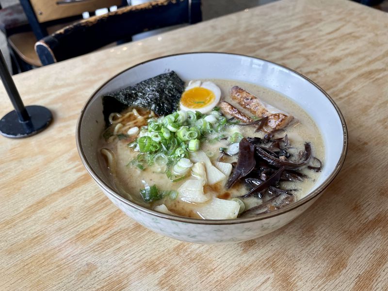 A white bowl of tonkotsu ramen with pork, egg, mushrooms, and scallions sitting on a wooden table. 