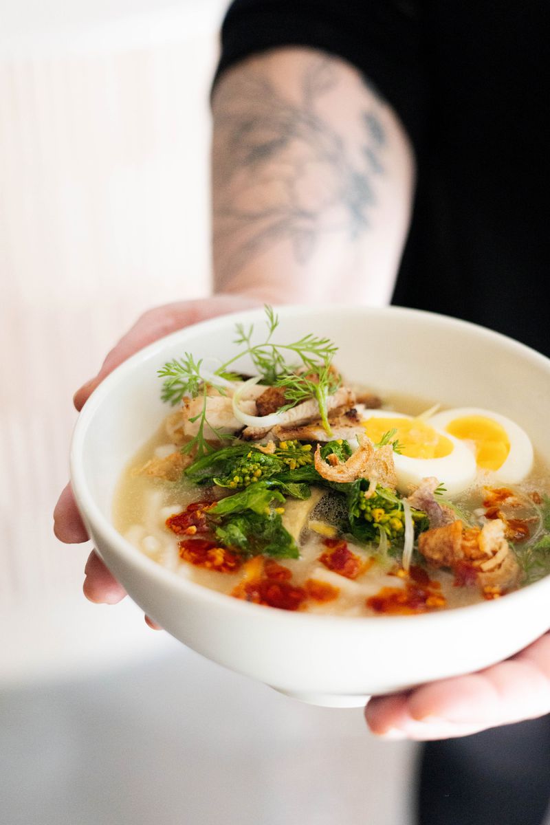 Two hands holding a white bowl of chicken noodle soup topped with greens, a halved boiled egg, and spicy chili sauce. 