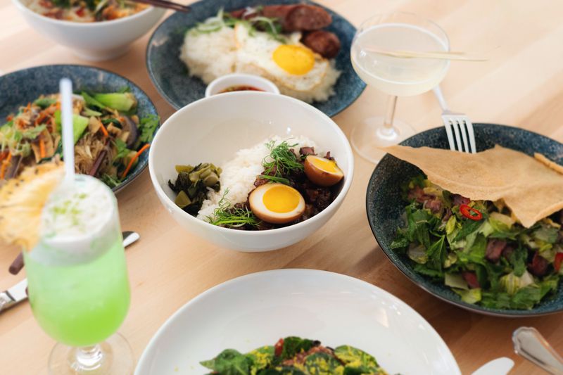 An assortment of dark blue and white dishes on a wooden table with cocktails interspersed among them, holding sweet pork and sticky rice with greens, and other food. 