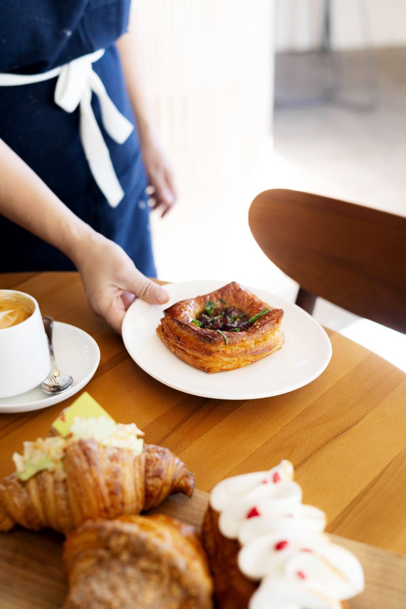 A hand placing a small white plate with a sweet pork Danish onto a wooden table that has other pastries and a cup of coffee sitting on it. 